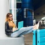 young girl with long hair black glasses is sitting outside airport she wears jeans black jacket jellow shoes she put her legs suitcase near she is bored typing laptop 1